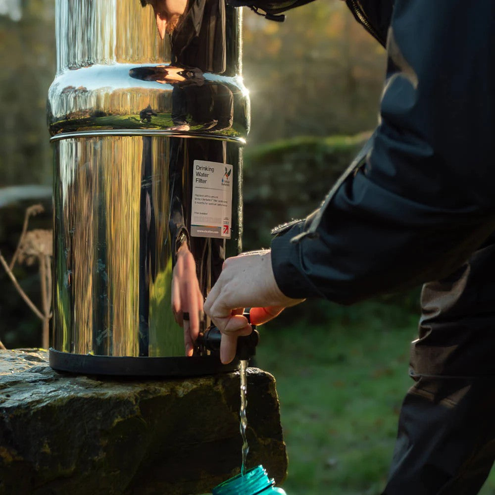 Person dispensing water from a polished stainless steel gravity water filter into a container outdoors, suitable for camping, emergency supplies, and farm use.