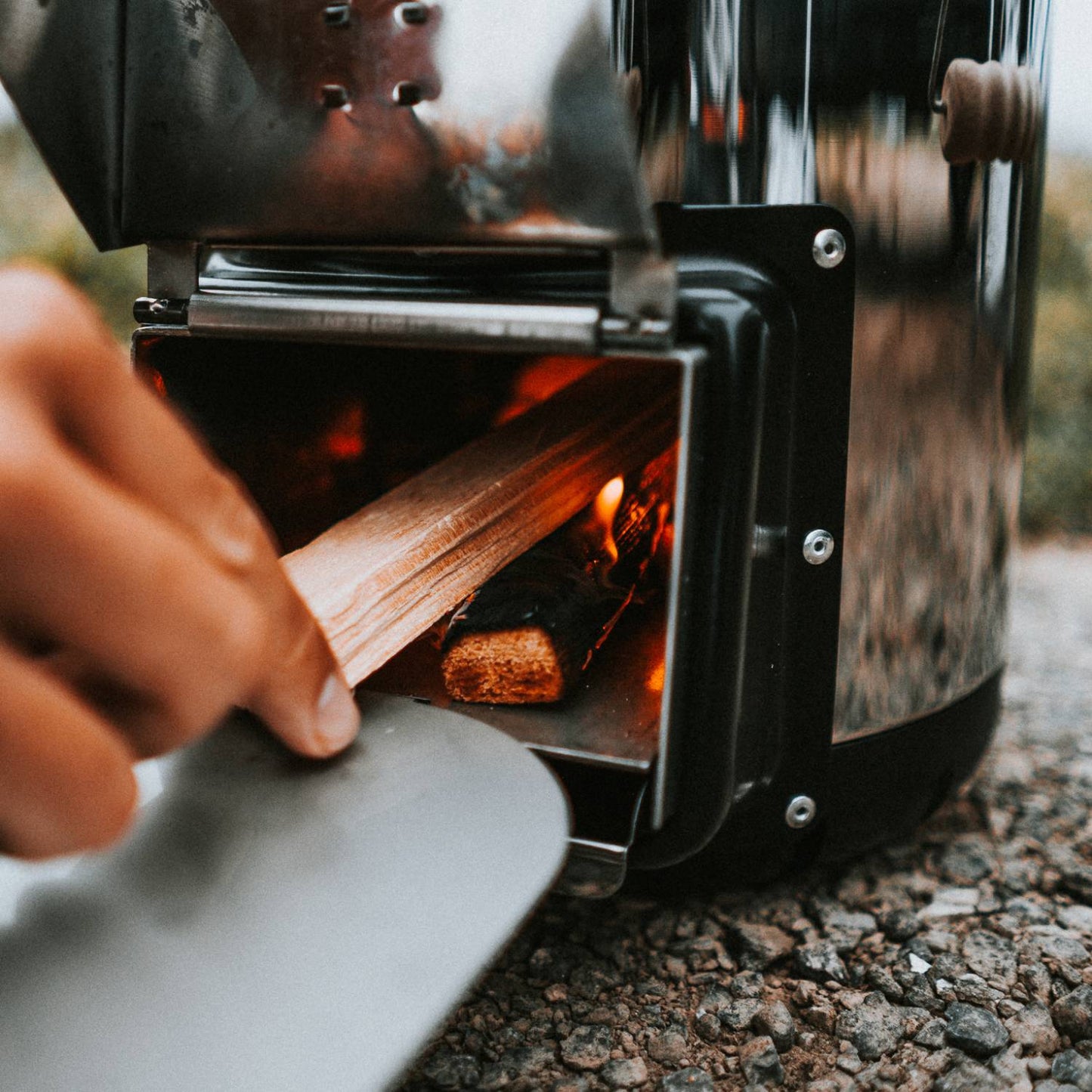 Hand placing kindling wood into the firebox of a Petromax Rocket Compact Outdoor Stove on a gravel surface, designed for quick heating in Farm, Garden & Tool, Camping, and Best Sellers collections.