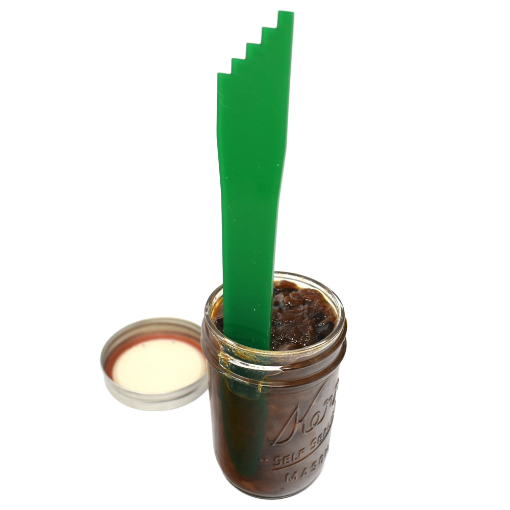 Green plastic canning bubble popper tool standing upright inside a glass jar filled with dark preserves, next to an open metal jar lid, for Kitchen & Food and Food Preservation collections.