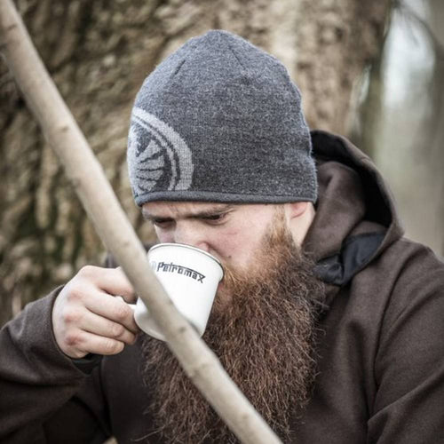 Man wearing a gray knit hat and brown hoodie sipping from a white enamel mug with black logo outdoors near a tree, suitable for Farm, Garden & Tool, Camping, and Brewing Coffee and Tea collections.