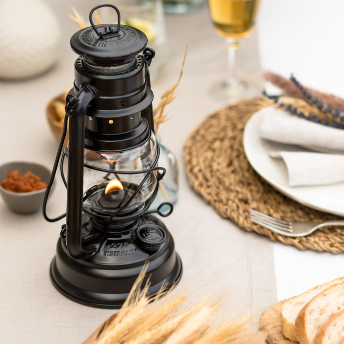 Handcrafted black galvanized oil lantern with thermal heat-resistant SUPRAX globe, softly glowing on a beige tablecloth beside rustic bread and elegant glassware, ideal for camping and outdoor use in lamps & lighting, emergency supplies, and camping collections.