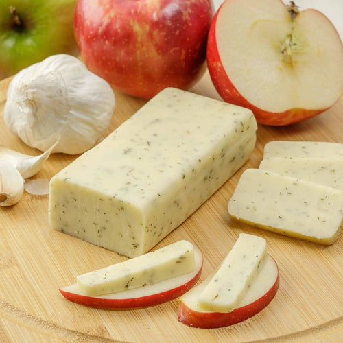 Handcrafted shelf-stable cheese block with herb flecks, served sliced on apple wedges with garlic cloves nearby, ideal for Kitchen & Food, Snacks and Sweets, Pantry, USA Made, Best Sellers, Promo Eligible collections.