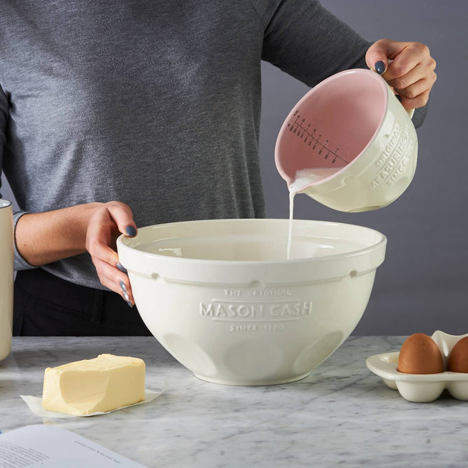Cream-colored measuring jug with pink interior gently pouring liquid into matching mixing bowl on marble countertop, surrounded by butter and eggs, featured in Kitchen & Food, Cookware, Baking Supplies, Food Prep, Measuring, Best Sellers, Promo Eligible collections.
