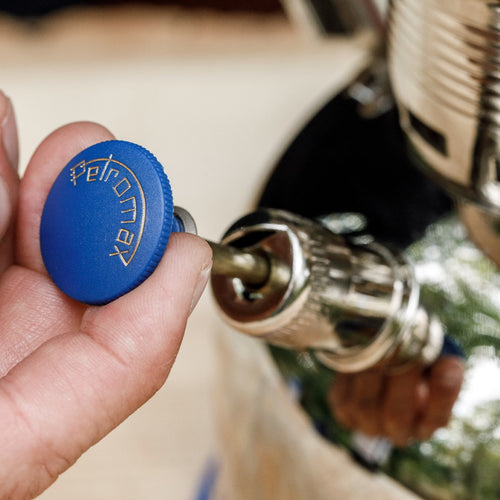 Hand gripping a brass and chrome pump component with a blue knob engraved with Petromax, designed for lamps, featured in Lamps & Lighting, Parts and Accessories, Other Parts, Best Sellers, and Promo Eligible collections.