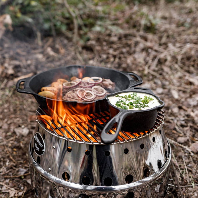Outdoor cooking scene with a cast iron skillet grilling sausages and onions alongside a small cast iron pot with creamy sauce garnished with herbs on a portable fire stove, ideal for Farm, Garden & Tool, Kitchen & Food, Camping, Cookware, Cast Iron, Best Sellers, and Promo Eligible collections.