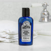 Blue glass bottle of unscented pre-shave oil with black cap and vintage-style label, set on a bathroom counter near folded white towels and a chrome faucet, showcasing a handcrafted grooming essential in Health & Wellness and USA Made collections.