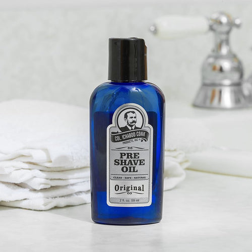 Blue glass bottle of unscented pre-shave oil with black cap and vintage-style label, set on a bathroom counter near folded white towels and a chrome faucet, showcasing a handcrafted grooming essential in Health & Wellness and USA Made collections.