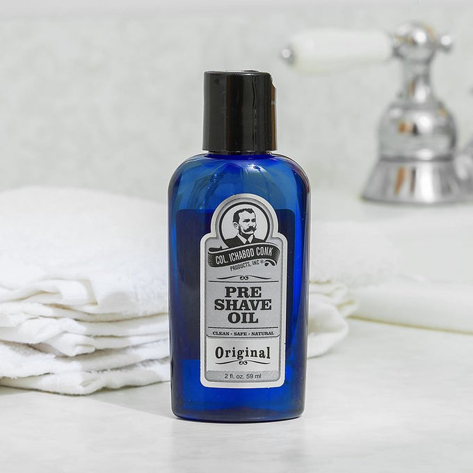 Blue glass bottle of unscented pre-shave oil with black cap and vintage-style label, set on a bathroom counter near folded white towels and a chrome faucet, showcasing a handcrafted grooming essential in Health & Wellness and USA Made collections.