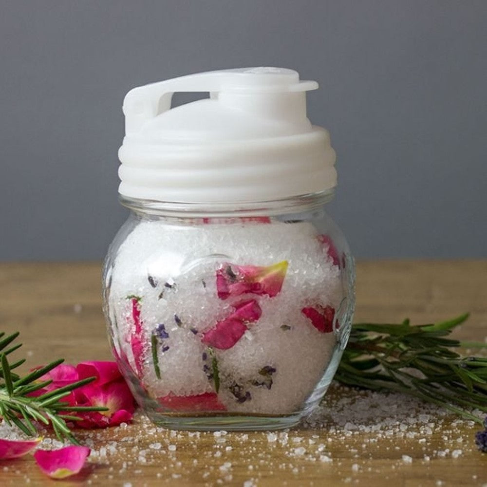 Clear glass jar with a white durable pour lid featuring a flip-top spout and carry loop, filled with coarse salt and pink flower petals, set among fresh herbs on a wooden surface, ideal for Kitchen & Food, Dinnerware, Kitchen Gadgets, Kitchen Accessories, USA Made, Best Sellers, and Promo Eligible collections.