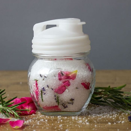 Clear glass jar with a white durable pour lid featuring a flip-top spout and carry loop, filled with coarse salt and pink flower petals, set among fresh herbs on a wooden surface, ideal for Kitchen & Food, Dinnerware, Kitchen Gadgets, Kitchen Accessories, USA Made, Best Sellers, and Promo Eligible collections.