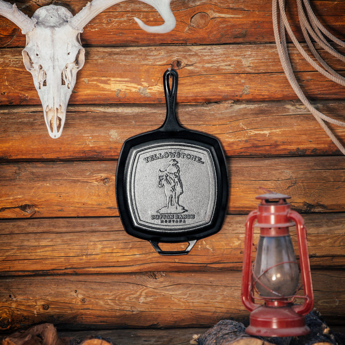 Handcrafted cast iron square grill pan with embossed cowboy design, made in USA, displayed against a rustic log wall with skull and vintage lantern, perfect for Kitchen & Food and Cast Iron collections.
