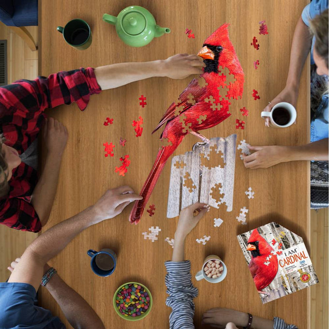 Four people assembling a 300-piece cardinal-shaped jigsaw puzzle on a wooden table, surrounded by mugs, a green teapot, and a bowl of colorful candy, featured in Toys, Books & Games and Puzzles collections.