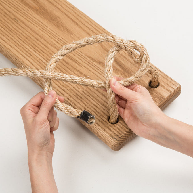 Close-up of hands threading sturdy natural rope through pre-drilled holes of a handmade wooden swing seat, crafted in USA, part of outdoor wooden toys and children's play collections.