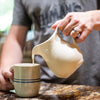 Handcrafted cream stoneware pitcher pouring liquid into a blue-striped ceramic mug, made in the USA, featured in Kitchen & Food, Dinnerware, Storage and Serving, USA Made, and Best Sellers collections.