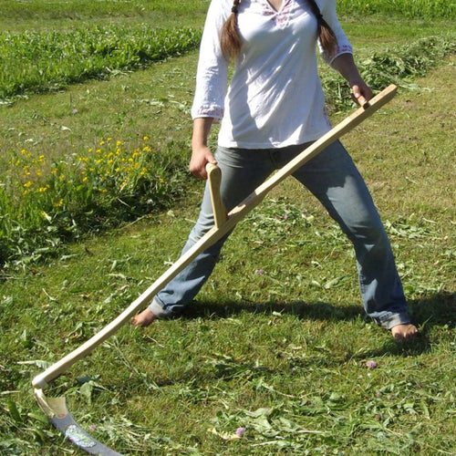 Person using a handcrafted wooden farming scythe with a curved metal blade to mow grass barefoot in a green garden, featured in Farm, Garden & Tool, Lawncare, Best Sellers, Promo Eligible collections.