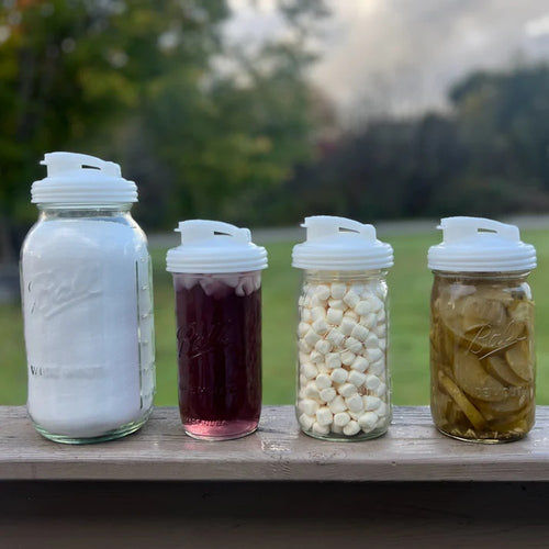Four clear wide-mouth glass jars with white pour lids featuring flip-top spouts and carry loops, filled with sugar, iced tea, mini marshmallows, and pickles, shown outdoors, ideal for Kitchen & Food, Dinnerware, Kitchen Gadgets, Kitchen Accessories, USA Made, and Best Sellers collections.