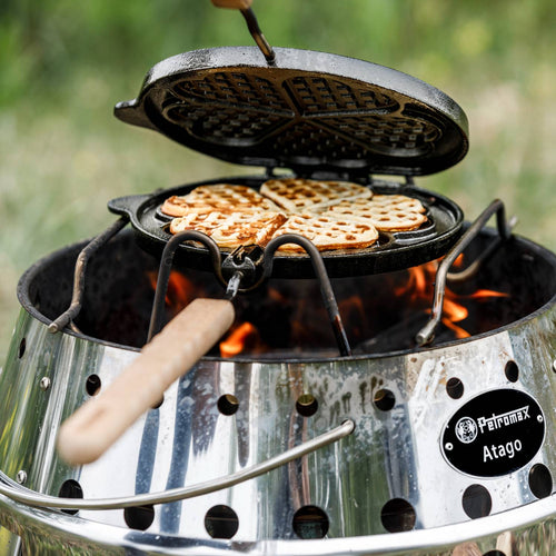 Cast iron rotating waffle iron with wooden handle cooking heart-shaped waffles over a campfire on a stainless steel fire pit, ideal for Farm, Garden & Tool and Camping collections.