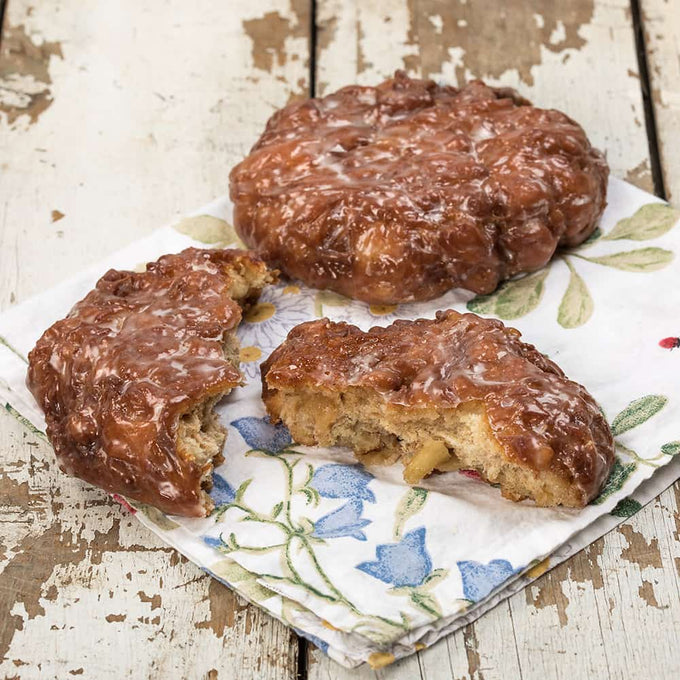 Two glazed fruit fritters, one whole and one broken to show fried fruit chunks inside, resting on a floral napkin on a rustic wooden surface, handcrafted and made in USA, ideal for Kitchen & Food, Snacks and Sweets, Holiday Hosting, USA Made, Best Sellers, Promo Eligible collections.