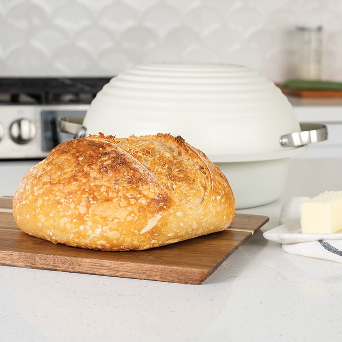 Golden crusted sourdough loaf resting on a wooden board next to a cream-colored baking cloche with metal handles, ideal for Kitchen & Food and Baking Supplies collections.