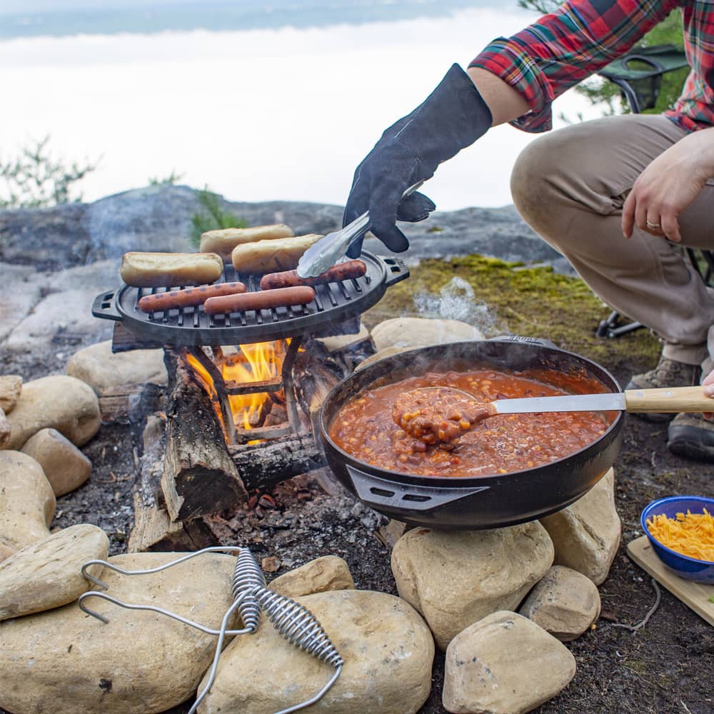 Handcrafted cast iron outdoor cookware featuring a two-piece set with a grilling grate cooking hot dogs and buns over a campfire, and a deep pan simmering chili, perfect for Camping, Kitchen & Food, Cast Iron, USA Made, Best Sellers, and Farm, Garden & Tool collections.