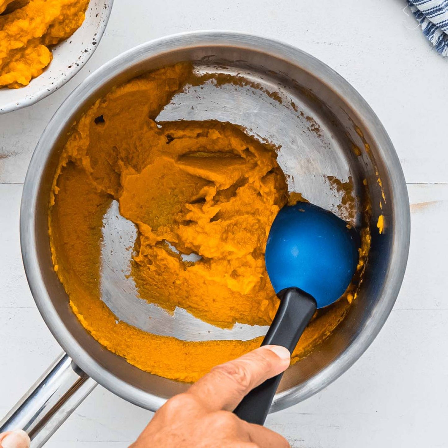 Hand using a flexible blue and black silicone spoon to scrape orange puree from a stainless steel saucepan, highlighting a versatile tool for Kitchen & Food, Food Prep, and Cooking Helpers collections.