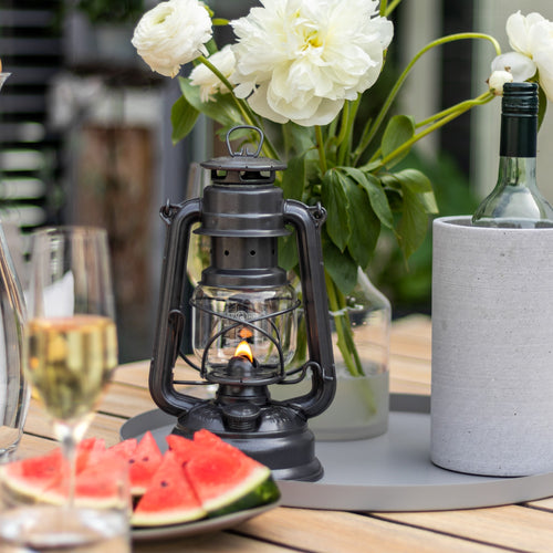 Handcrafted galvanized oil lantern with a warm glowing flame, displayed on a wooden table with fresh white flowers, watermelon slices, wine glass, and bottle cooler, ideal for outdoor lighting and camping collections.