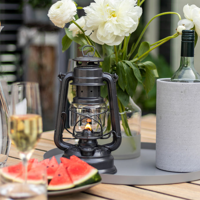 Handcrafted galvanized oil lantern with a warm glowing flame, displayed on a wooden table with fresh white flowers, watermelon slices, wine glass, and bottle cooler, ideal for outdoor lighting and camping collections.