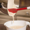 Red silicone batter spatula with slotted flexible head and sturdy white plastic handle scraping thick batter from a stainless steel mixing bowl into a white bowl, featured in Kitchen & Food, Kitchen Gadgets, Cooking and Baking Helpers, Best Sellers, and Promo Eligible collections.