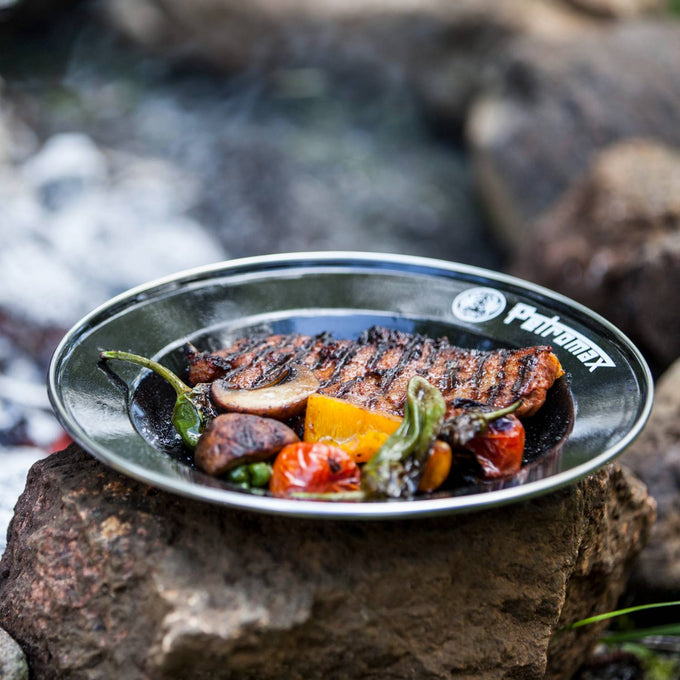 Black enamel plate with Petromax logo holding grilled steak and charred vegetables, placed on a rock near a campfire, ideal for Farm, Garden & Tool, Kitchen & Food, Camping, Dinnerware, and Best Sellers collections.