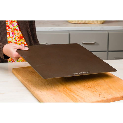 Hand holding a slim, dark baking steel above a wooden cutting board in a kitchen setting, ideal for Kitchen & Food, Cookware, Baking Supplies, and Bread Making collections.