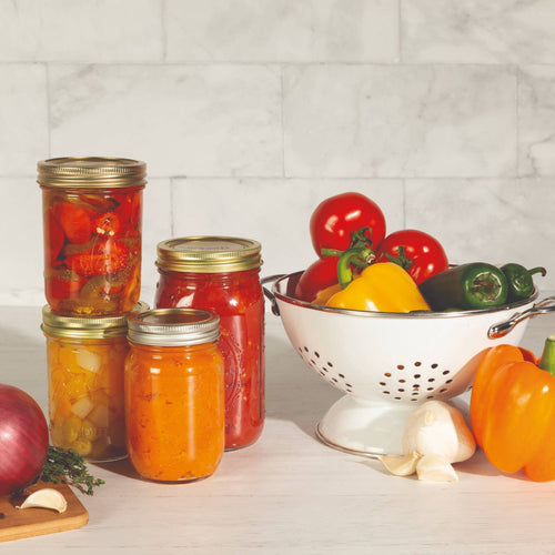 Four glass jars sealed with gold-tone wide-mouth canning lids filled with preserved vegetables and sauces, placed beside fresh colorful peppers and garlic in a white metal colander, suitable for Kitchen & Food, Food Preservation, and Canning collections.