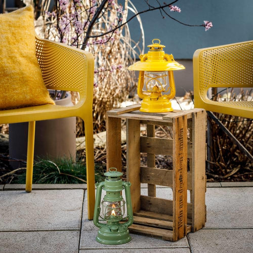 Handcrafted green and yellow galvanized oil lanterns with thermal heat-resistant globes, one lit and placed on a rustic wooden crate between yellow modern chairs, ideal for Lamps & Lighting, Camping, and Emergency Supplies collections.