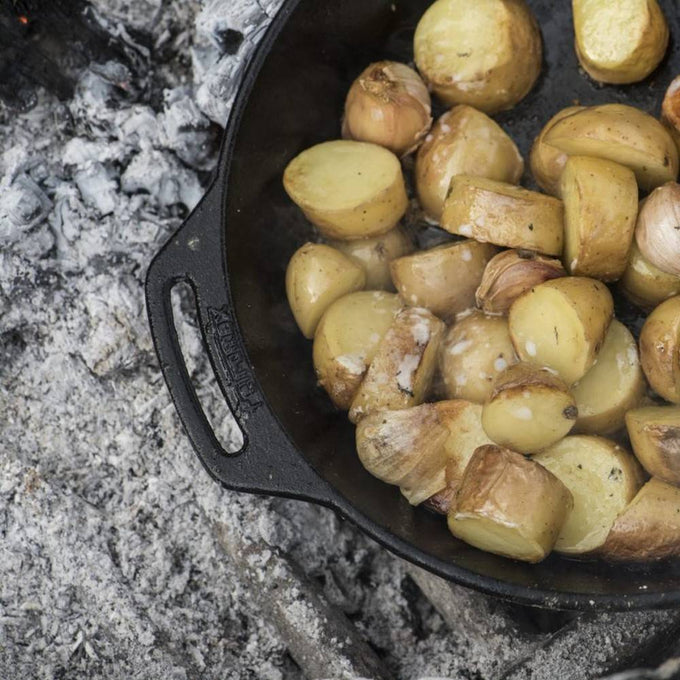 Cast iron skillet with two side handles cooking halved potatoes over hot embers, showcasing durable cookware ideal for farm, garden, camping, and kitchen use.