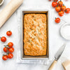 Large loaf baking in a recycled steel pan with corrugated texture for even baking, set on parchment with fresh cherry tomatoes, a rolling pin, and a knife nearby in a bright kitchen setting.