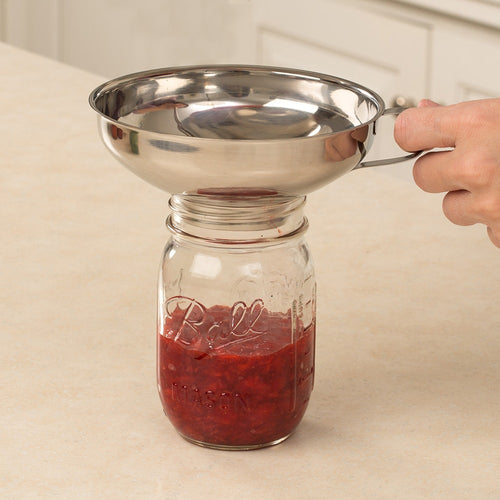 Stainless steel funnel held over a Ball® canning jar filled with red fruit preserves on a kitchen counter, part of food preservation and canning essentials.