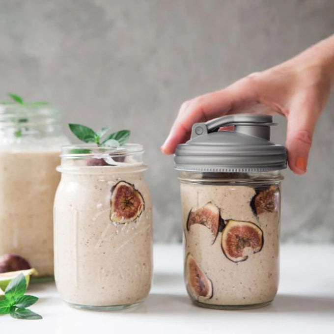 Hand placing a gray plastic pour lid with carry loop on a regular-mouth glass jar filled with creamy mixture and dried fig slices, showcasing USA-made kitchen accessories for Kitchen Gadgets and Best Sellers collections.