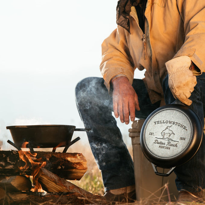 Person in outdoor gear holding a cast iron cookware piece with a Bucking Bronco design next to a wood fire cooking setup, featuring kitchen and camping themes.