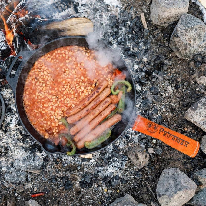 Cast iron skillet with long handle cooking sausages, baked beans, and peppers over an open campfire surrounded by rocks, ideal for Farm, Garden & Tool, Kitchen & Food, Camping, Cookware, Cast Iron, Best Sellers, and Promo Eligible collections.