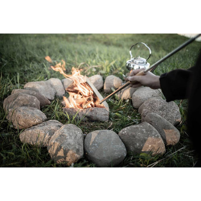 Hand holding a long metal fire blow pipe directing air into a small campfire surrounded by a circle of rocks outdoors, suitable for camping and hearth accessories in Farm, Garden & Tool collections.