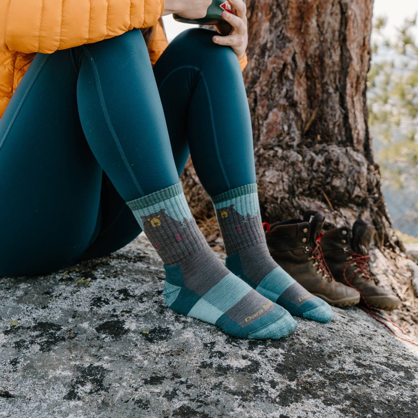 Woman wearing green and gray micro crew socks with bear design, seated on a rock near hiking boots and tree trunk, highlighting breathable lightweight comfort in outdoor setting for Health & Wellness, Camping, USA Made, and Best Sellers collections.