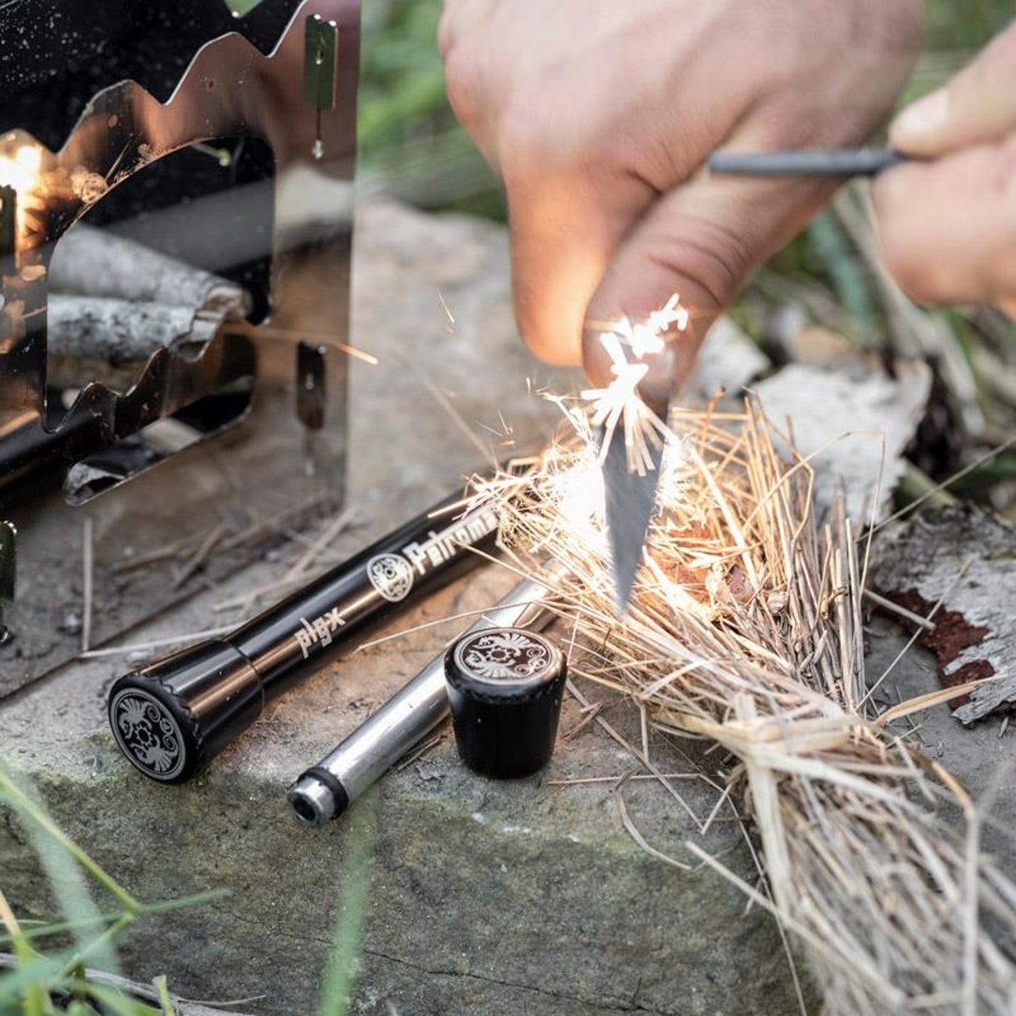Hand striking a steel rod to create sparks on dry tinder beside two black fire plungers and a small black container, set outdoors for camping or emergency use.
