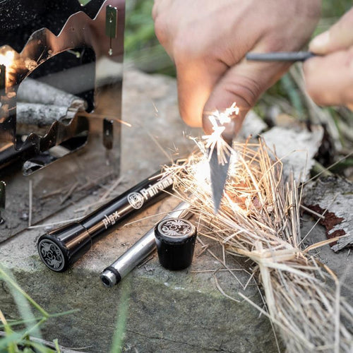 Hand striking a steel rod to create sparks on dry tinder beside two black fire plungers and a small black container, set outdoors for camping or emergency use.