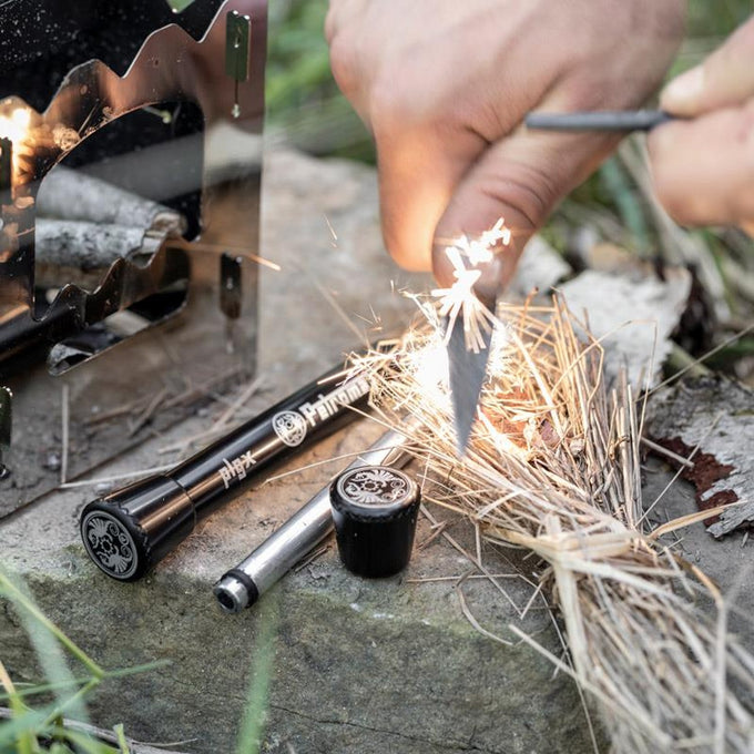 Hand striking a steel rod to create sparks on dry tinder beside two black fire plungers and a small black container, set outdoors for camping or emergency use.