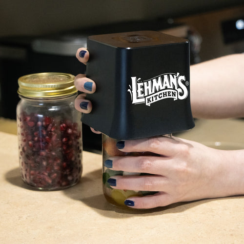 Hands holding a black cordless vacuum sealer device securing a glass mason jar filled with preserved food, alongside another jar of pomegranate seeds on a kitchen countertop, featured in Kitchen & Food and Kitchen Gadgets collections.