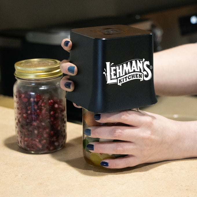 Hands holding a black cordless vacuum sealer device securing a glass mason jar filled with preserved food, alongside another jar of pomegranate seeds on a kitchen countertop, featured in Kitchen & Food and Kitchen Gadgets collections.