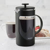 Black plastic French press coffee maker with a stainless steel filter rod, filled with dark brewed coffee, shown on a kitchen counter next to a glass cup of coffee and a plate of cookies, ideal for Kitchen & Food and Brewing Coffee and Tea collections.