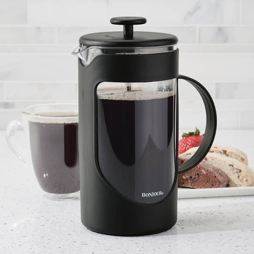 Black plastic French press coffee maker with a stainless steel filter rod, filled with dark brewed coffee, shown on a kitchen counter next to a glass cup of coffee and a plate of cookies, ideal for Kitchen & Food and Brewing Coffee and Tea collections.