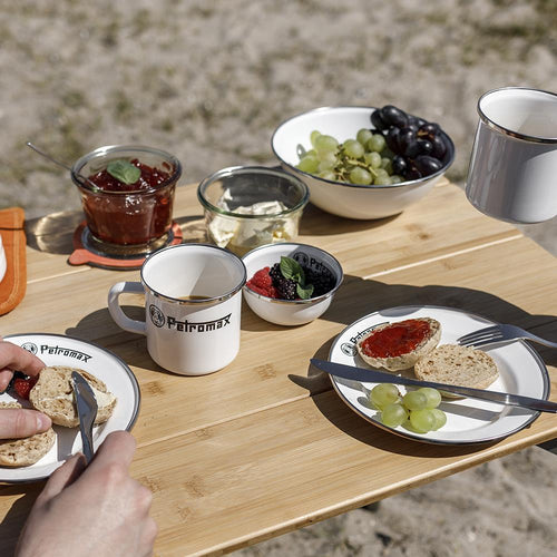 Two enamel plates with black rims and logo on a wooden tray outdoors, holding a bread roll with jam and green grapes, accompanied by matching mugs and bowls with fruit, ideal for Farm, Garden & Tool, Kitchen & Food, Camping, Dinnerware, Best Sellers, Promo Eligible collections.
