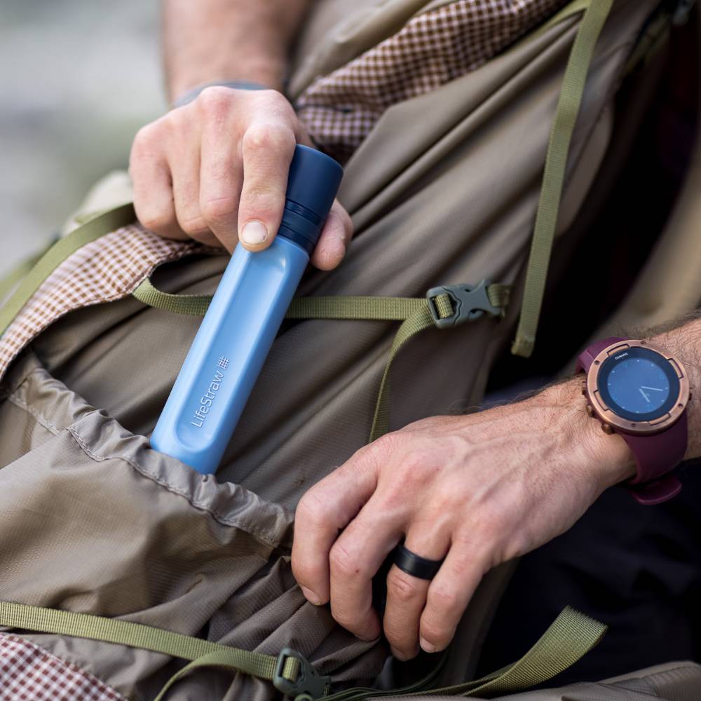 Person placing a blue water filter straw into a spacious pocket of a brown outdoor backpack, showcasing a portable survival tool for camping and emergency supplies collections.
