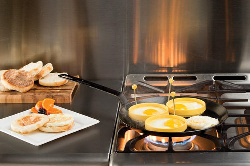 Seasoned steel skillet on gas stove cooking eggs in yellow silicone rings, with toasted English muffins on a white plate and cutting board nearby, ideal for Kitchen & Food, Cookware, Pots and Pans, USA Made, Best Sellers, and Promo Eligible collections.